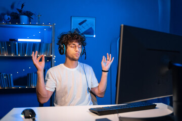 Arabic guy gamer meditates sitting before computer in a headset at night in neon light, closed his eyes making yoga.