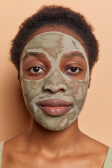 Headshot of serious African woman immerses herself in self care applying clay mask with serene expression emphasizing importance of skincare rituals and rejuvenating benefits poses indoor brown wall