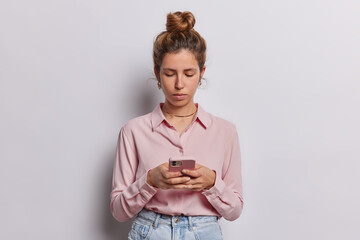 Horizontal shot of serious attentive European woman with piercing in nose uses mobile phone for chatting online sends text messages wears formal shirt and jeans isolated over white background.