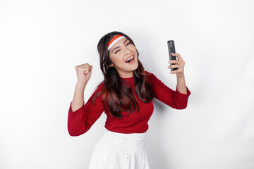 A young Asian woman with a happy successful expression while holding her phone and wearing red top...