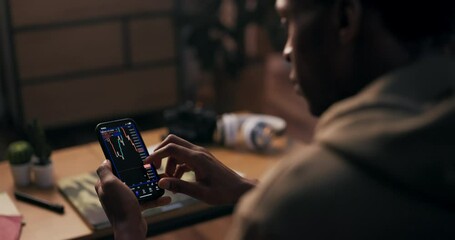 Active Day Trader man sitting on a couch, engrossed in a trading app on his cellphone, making real-time investment decisions.