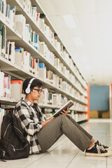 Naklejka premium Young hipster student sitting on the floor with his back against a bookcase full of books studying or doing research and listening to music in headphones.