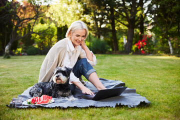 Mature woman using laptop in backyard garden sitting on blanket with her dog