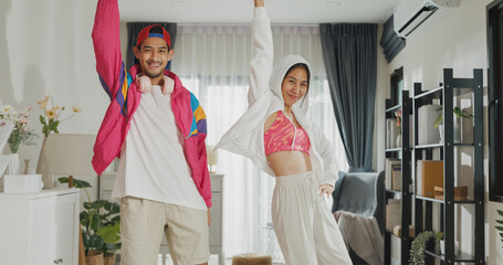 Happy young Asian funny influencer couple joyful looking at camera enjoy dancing trendy music together in living room at home on holiday. Smile young husband and wife moving for favorite song.
