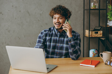 Successful young employee business Indian man wear casual blue checkered shirt talk speak on mobile...