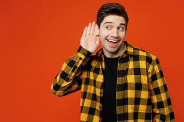 Young curious nosy caucasian man he wears yellow checkered shirt black t-shirt try to hear you overhear listening intently isolated on plain red orange background studio portrait. Lifestyle concept.