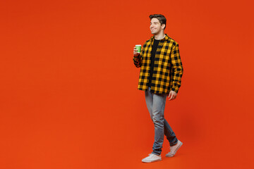 Full body side view young caucasian man he wear yellow checkered shirt black t-shirt hold takeaway delivery craft paper brown cup coffee to go isolated on plain red orange background studio portrait.