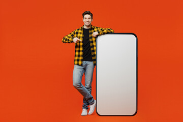 Full body young man he wear yellow checkered shirt black t-shirt point index finger on big huge blank screen mobile cell phone smartphone with workspace area isolated on plain red orange background.