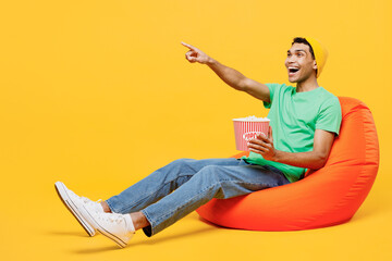 Full body young man of African American ethnicity he wears casual clothes green t-shirt hat sit in bag chair eat popcorn point index finger aside watch movie film isolated on plain yellow background.