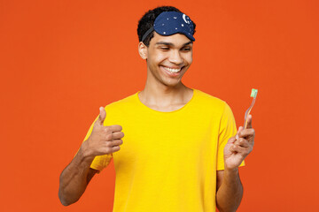 Calm smiling young man wear pyjamas jam sleep eye mask rest relax at home brushing teeth show thumb up isolated on plain orange background studio portrait. Good mood night nap, daily routine concept. - Powered by Adobe