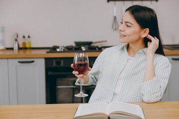 Side view minded fun happy smiling young happy housewife latin woman wear casual clothes striped shirt drink wine look aside sit at table in light kitchen at home alone Lifestyle cooking food concept