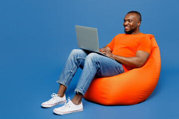 Full body young IT man of African American ethnicity he wear orange t-shirt sit in bag chair hold...