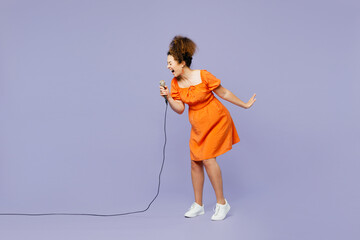 Full body side view young expressive latin woman wear orange blouse casual clothes sing song in microphone at karaoke club isolated on plain pastel light purple background studio. Lifestyle concept - obrazy, fototapety, plakaty