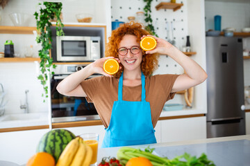 In a modern kitchen, a red-haired woman holds orange slices near her face. With a smile, she...