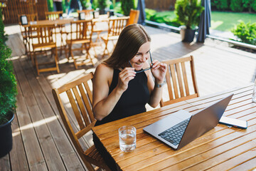 Young cute woman, freelancer, developer, enjoying coffee break on summer terrace of cozy cafe, working remotely on laptop, smiling looking at laptop