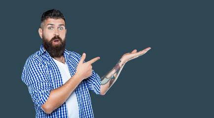 surprised bearded man wear checkered shirt presenting product on grey background with copy space