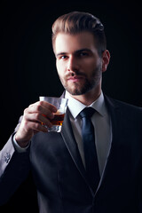 Portrait of serious elegant handsome young man in suit toasting with glass of alcohol isolated on black background.