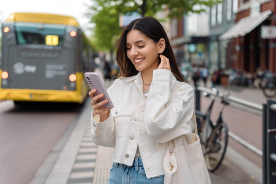 Happy Commute On Public Transport. Multi-ethnic Attractive Woman With Beautiful Smile At Bus Station Looking At Phone