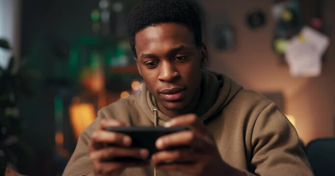 African man sits on a couch in his living room, clutching his cellphone tightly with a look of frustration on his face. He plays a challenging mobile game that causing him stress and disappointment.