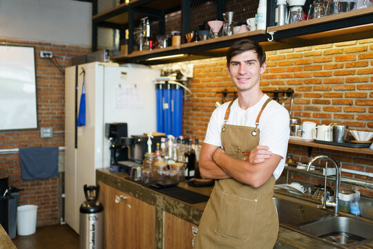 Professional young coffee barista standing inside the counter bar in the coffee shop and making arms crossed and looking at camera, handsome - good looking caucasian ethnic barista posing.