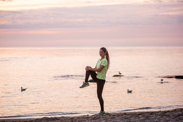 Fototapeta na wymiar Young woman in sportswear is warming up early in morning on seashore at sunrise. Girl of slim build with long hair is stretching. Outdoor sports by the water. Fitness classes, body care, mental health