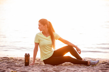 Fototapeta na wymiar Young woman in sportswear is sitting on the seashore at sunrise. Girl is resting after workout, replenishes her water balance, drinks from a sports bottle, looks at the water. Morning outdoor fitness 
