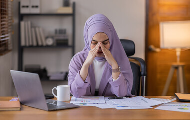 A stressed Asian business asian islamic woman in hijab holding documents is working from home, feeling stressed because of lot of paperwork appearing worried, tired, and overwhelmed