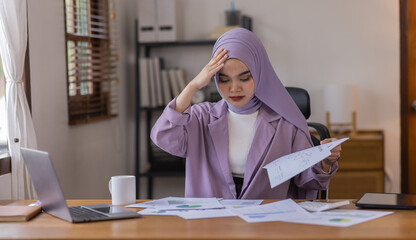 A stressed Asian business asian islamic woman in hijab holding documents is working from home, feeling stressed because of lot of paperwork appearing worried, tired, and overwhelmed