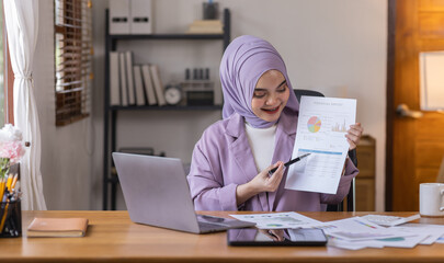 Fototapeta na wymiar Asian business asian islamic woman in hijab holding financial documents to check calculations on laptop or analyzing report accuracy to show data with graphs to prepare company finances