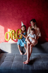 Young mother sits on sofa with small children near red wall on gray sofa. Family with one parent celebrates the first year of baby. Older sister is holding foil balloons ONE. Lifestyle party at home.