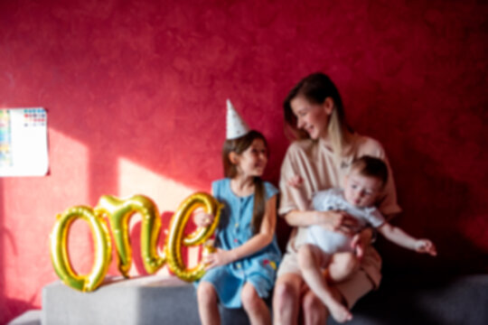 Defocused Mother is fooling around on sofa with small children near red wall on gray sofa. Family with one parent celebrates babys first year. Older sister is holding foil balloons ONE. Lifestyle
