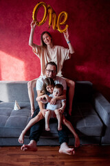 Obraz na płótnie Canvas Portrait of happy, diversity family on the birthday of one year old boy. Mother laughs, holds foil balloons ONE in hands, father hugs daughter and son on gray sofa in the sun. Home holiday party.