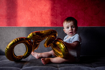 First birthday party, baby plays with gold colored foil balloons ONE. Little boy fooling around,...