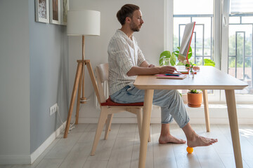Man freelancer working on computer at home doing foot exercise using massage ball under desk for...