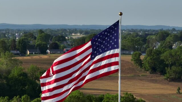 Big iconic American flag swaying, picturesque countryside in background, aerial orbit. Long drone zoom lens.