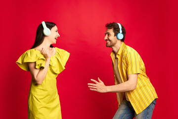 Positive young man and woman in bright yellow clothes listening to music in headphones and dancing over red studio background. Concept of friendship, relationship, communication, emotions, lifestyle