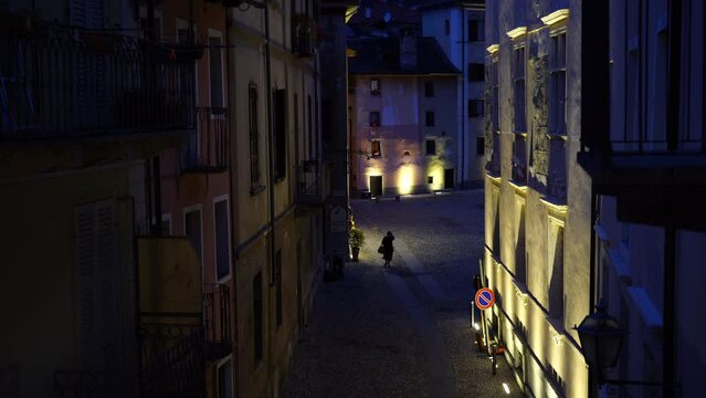 An anonymous woman walking in a street in the old town of Domodossola, Italy, at night.