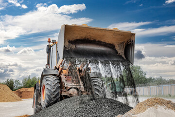 Powerful wheel loader or bulldozer on sky background. Loader pours crushed stone or gravel from the bucket. Powerful modern equipment for earthworks and bulk handling.