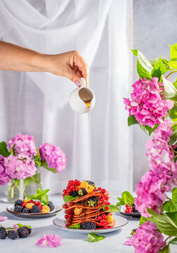 Women hand is pouring syrup from jar to pink pancake with berry and mint in ceramic dish surrounded pink hydrangea blossom