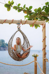 Smiling caucasian tanned girl with blond hair sitting on wicker swing in lotus position on beach. Relaxed girl sits on swing during her summer vacation on tropical beach. Happy tourist concept