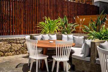 Open-air relaxation area with  brown wooden round table. Surrounding the table is a semi-round sofa. Three elegant white chairs complement the setting, offering additional seating options.