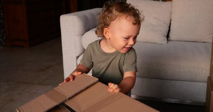 Cheerful toddler happily plays with cardboard boxes, smiling and enjoying fun. Slow motion footage captures joy and emotions on child's face. Young boy engagement with unfamiliar objects