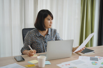 Asian business woman working on laptop and paper documents, holding pen in the office.