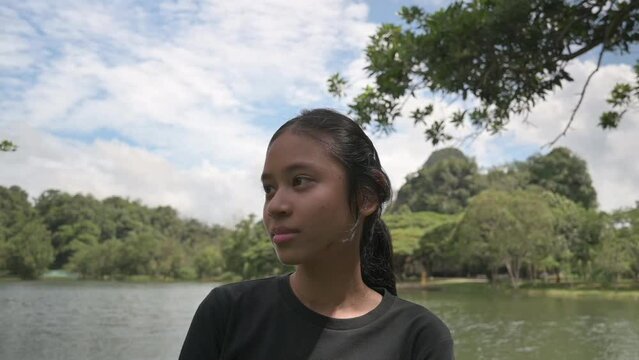 Shy adorable girl filming a video under the shade of the tree in nature park during summer. Portrait embarrassed female teenager smiling and looking at camera while standing near the lagoon. 