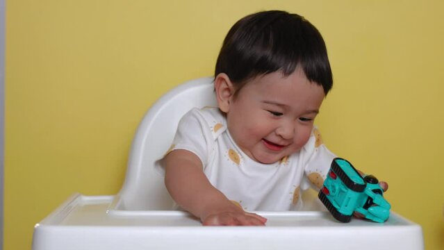Cute asian baby sits on highchair with toy and smiling on yellow background