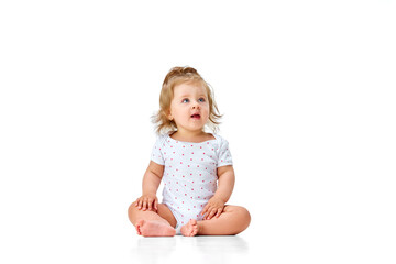 Curious child. Little, cute baby girl, toddler in diaper sitting on floor and attentively looking against white studio background