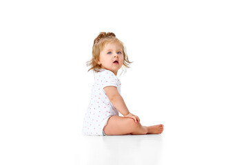 Curious child. Little, cute baby girl, toddler in diaper sitting on floor and attentively looking against white studio background
