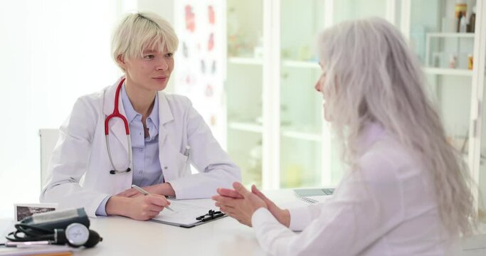 Female doctor listens, elderly woman talks about chronic diseases health complaints symptoms of ailment. Therapist takes notes fills out history form during patient visit