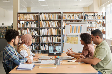 Group of foreign students having online lesson with teacher while sitting in the library