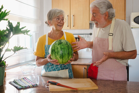 Happy Senior Caucasian Couple In Home Kitchen Holding A Big And Heavy Seasonal Watermelon - Hydration, Freshness, Diet And Healthy Eating Lifestyle Concept